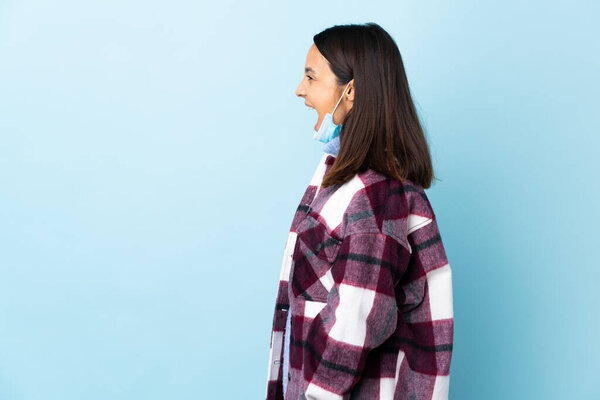 Young brunette mixed race woman protecting from the coronavirus with a mask and gloves over isolated blue background laughing in lateral position