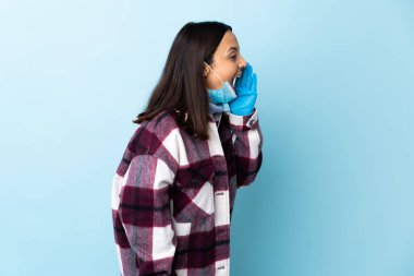 Genç, esmer, melez bir kadın. Koronavirüsten koruyor. Maske ve eldivenlerle izole edilmiş mavi arka planda.