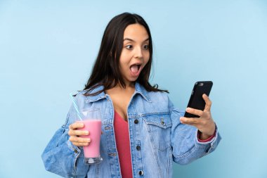 İzole edilmiş mavi arka planda çilekli milkshake 'i olan genç bir kadın elinde kahve ve bir cep telefonu tutuyor.