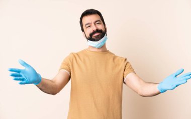 Koronavirüsten sakallı beyaz bir adam maske ve eldivenlerle korunuyor. İzole bir arka plan sunuyor ve davet ediyor.