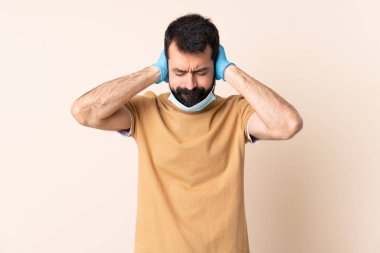 Koronavirüsten sakallı, maskeli ve eldivenli beyaz bir adam izole edilmiş arka planda hayal kırıklığına uğramış ve kulaklarını kapatmış.