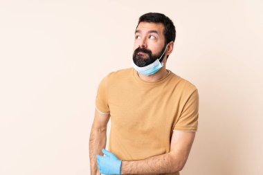 Sakallı, maskeli ve eldivenli beyaz bir adam omuzlarını kaldırırken şüpheli hareketlerde bulunuyor.