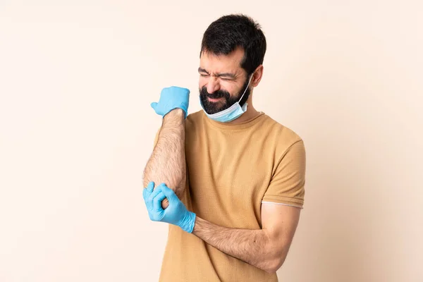 髭を生やした白人男性は 肘の痛みで隔離された背景の上にマスクと手袋でコロナウイルスから保護します — ストック写真