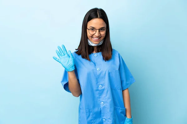 在孤立的蓝色背景上的外科医生妇女 带着愉快的表情用手向她致意 — 图库照片