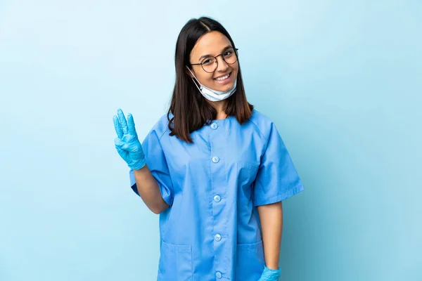 在孤立的蓝色背景上的外科医生女人快乐地数着三个手指 — 图库照片