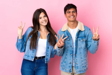Soyutlanmış pembe arka planda genç bir çift gülümsüyor ve iki eliyle zafer işareti gösteriyor.
