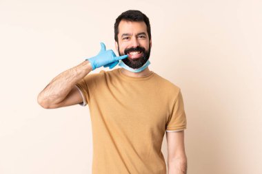 Koronavirüsten sakallı, maskeli ve eldivenli beyaz bir adam telefon jesti yapıyor. Bana geri dönüş işareti ver.