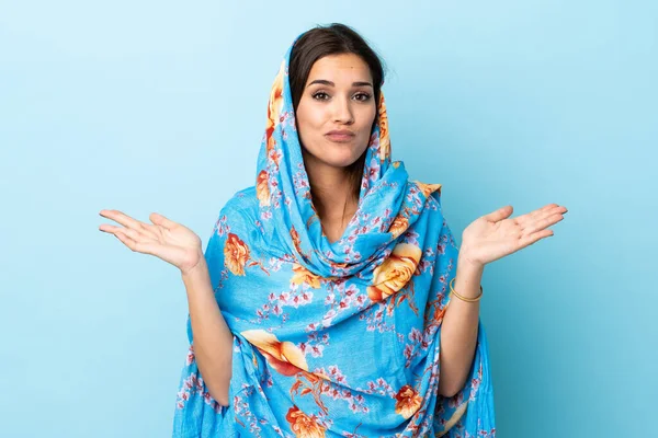 Young Moroccan woman with traditional costume isolated on blue background having doubts while raising hands