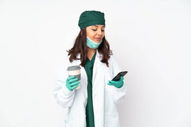 Yeşil üniformalı kadın, beyaz arka planda izole edilmiş. Elinde kahve ve cep telefonu var.