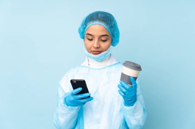Mavi üniformalı genç bir cerrah elinde kahve ve bir cep telefonu tutuyor.