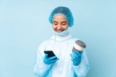 Mavi üniformalı genç bir cerrah elinde kahve ve bir cep telefonu tutuyor.