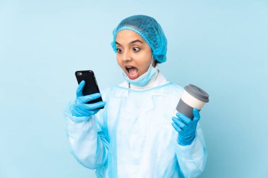 Mavi üniformalı genç bir cerrah elinde kahve ve bir cep telefonu tutuyor.