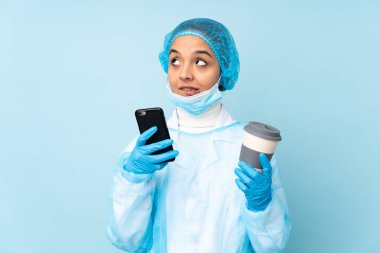 Mavi üniformalı genç bir cerrah elinde kahve ve bir şeyler düşünürken cep telefonu tutuyor.
