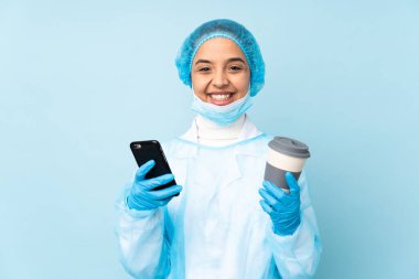 Mavi üniformalı genç bir cerrah elinde kahve ve bir cep telefonu tutuyor.