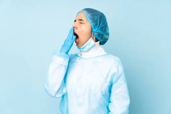 Young surgeon Indian woman in blue uniform yawning and covering wide open mouth with hand