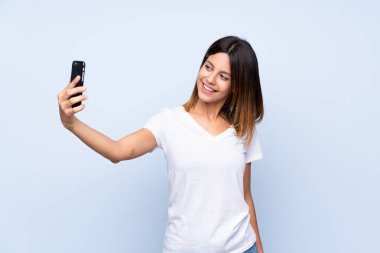 İzole edilmiş mavi arka planda genç bir kadın cep telefonuyla selfie çekiyor.