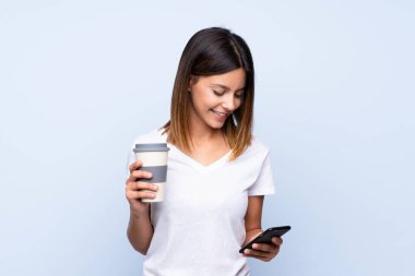 İzole edilmiş mavi arka planda, elinde kahve ve cep telefonu olan genç bir kadın.