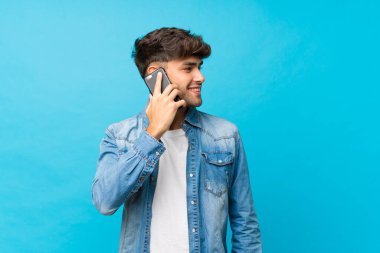 Soyutlanmış mavi arka planda genç yakışıklı bir adam cep telefonuyla sohbet ediyor.