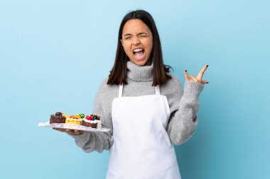 Pasta şefi mavi arka planda büyük bir pasta tutuyor ve boynuz hareketi yapıyor..