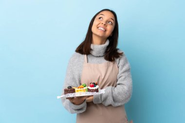 Pasta şefi mavi arka planda büyük bir pasta tutuyor gülümserken yukarı bakıyor..