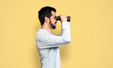 Handsome man with beard with binoculars over isolated yellow background