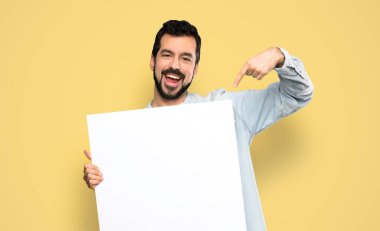 Handsome man with beard holding an empty placard over isolated yellow background