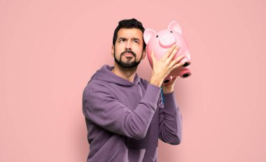 Handsome man with sweatshirt holding a piggybank over isolated pink background