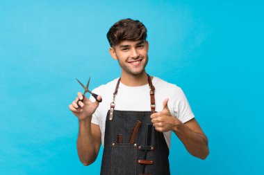 İzole edilmiş mavi geçmişi olan genç yakışıklı bir adam kuaför ya da başparmağı havada berber elbisesi