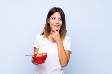 Young woman over isolated blue background holding a bowl of cereals and thinking
