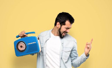 Handsome man with beard holding a radio over isolated yellow background