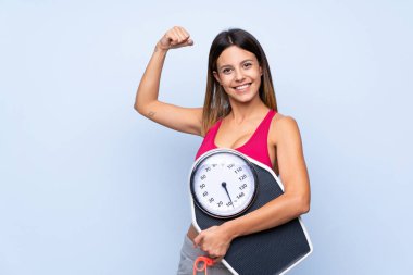 Tartı makinesi olan bir kız, izole edilmiş mavi arka planda elinde bir tartı makinesiyle güçlü bir hareket yapıyor.