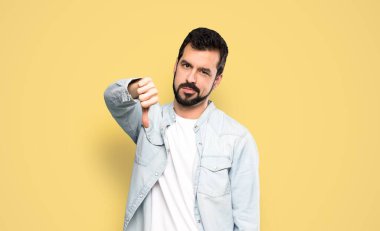 Sakallı yakışıklı adam, izole edilmiş sarı arka planda olumsuz bir ifadeyle baş parmağını aşağı gösteriyor.