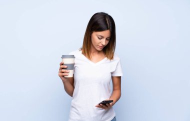 İzole edilmiş mavi arka planda, elinde kahve ve cep telefonu olan genç bir kadın.