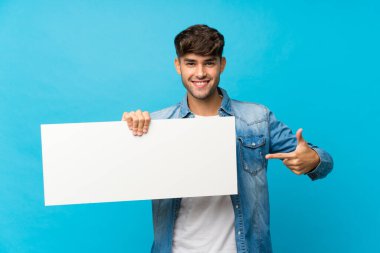 Soyutlanmış mavi arka plan üzerinde genç yakışıklı bir adam bir kavram eklemek için boş beyaz bir levha tutuyor.
