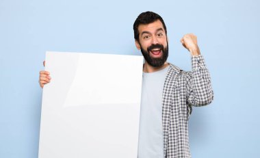 Happy Handsome man with beard holding an empty placard over isolated blue background