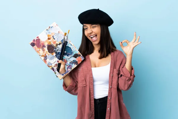 年轻的艺术家女人在孤立的蓝色背景上拿着一个调色板 用手指显示好的标志 — 图库照片