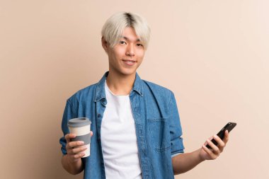 Arka planda kalan Asyalı genç bir adam kahve ve cep telefonu tutuyor.