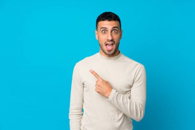 Soyutlanmış mavi arka planı olan genç adam şaşırmış ve işaret ediyor.