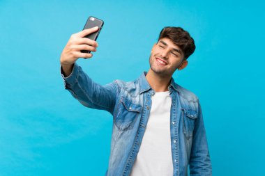 Soyutlanmış mavi arka planda genç yakışıklı bir adam cep telefonuyla selfie çekiyor.