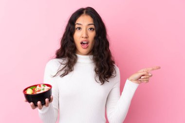 Karışık ırktan bir kadın bir kase erişteyi soyutlanmış pembe arka planda tutuyor. Şaşırmış ve işaret ediyor.