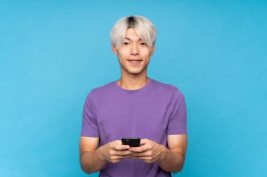 Asyalı genç adam izole edilmiş mavi arka planda cep telefonuyla mesaj gönderiyor.