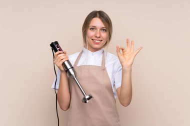 Genç sarışın kadın el blenderi kullanıyor. Parmaklarıyla işaret veriyor.