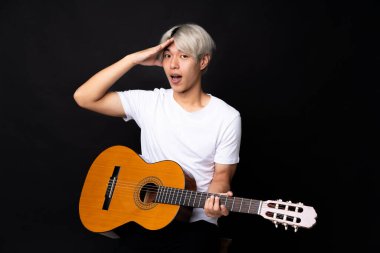 Siyah arka planda gitar çalan Asyalı genç bir adam şaşırmış ve şok olmuş bir yüz ifadesi var.