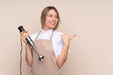 Genç sarışın kadın el blenderi kullanıyor. Yan tarafı göstererek bir ürün sunuyor.