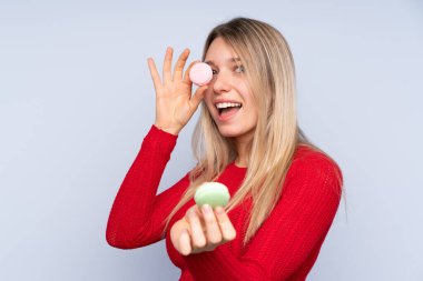 Soyutlanmış mavi arka planda genç sarışın kadın elinde renkli Fransız badem kurabiyeleri tutuyor.