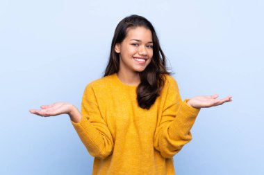 İzole edilmiş mavi arka planda süveteri olan genç Kolombiyalı kız iki eliyle kopyalama alanı tutuyor.