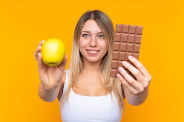 Bir elinde çikolata tableti, diğer elinde elma olan izole edilmiş mavi arka planda genç sarışın bir kadın.
