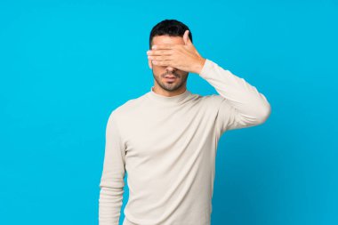 İzole edilmiş mavi arka planı olan genç bir adam gözleri elleriyle kapatıyor. Bir şey görmek istemiyorum.
