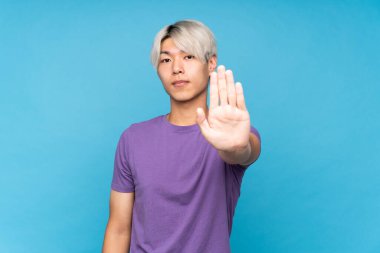 İzole edilmiş mavi arka planda genç Asyalı adam dur hareketi yapıyor.