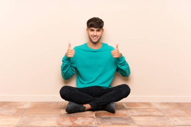 Genç yakışıklı bir adam yerde oturuyor ve baş parmağını kaldırıyor.